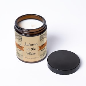 May include: A brown glass candle jar with a white wax fill and a black lid. The label features a beige background with orange pumpkin illustrations, black text reading Autumn in the Shire, and the brand name JOYFULCANDLESGB at the bottom.