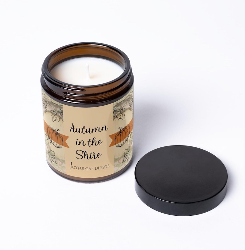 May include: A brown glass candle jar with a white wax fill and a black lid. The label features a beige background with orange pumpkin illustrations, black text reading Autumn in the Shire, and the brand name JOYFULCANDLESGB at the bottom.