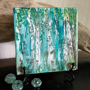 May include: A square decorative tile featuring an abstract painting of a birch forest. The artwork displays white tree trunks with dark markings against a vibrant background of teal, turquoise, and bright green foliage, rendered in a fluid style.