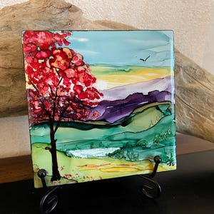 May include: Square artwork featuring a vibrant landscape with a red-leafed tree in the foreground. The background shows rolling hills in shades of green, purple, and yellow under a blue sky. The artwork is displayed on a black metal stand.