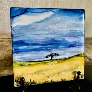 May include: A square ceramic tile featuring an alcohol ink landscape painting. The artwork displays a vibrant yellow field in the foreground, a single black silhouette of a tree in the center, and a layered blue and purple sky above the horizon.