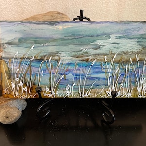 May include: A rectangular artwork depicting a coastal scene with a blue and teal sky, a blue body of water, and white seagrass. The artwork is mounted on a black metal easel. The painting is approximately 6 inches wide and 4 inches tall.