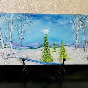 May include: A rectangular glass art piece depicting a winter landscape. The scene features snow-covered ground, evergreen trees, and birch trees under a blue sky with a full moon. The artwork is displayed on a black metal stand.