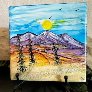 May include: Square artwork depicting a mountain landscape with a yellow sun, blue sky, and three black birds. The mountains are purple and white, with green and brown trees in the foreground. The artwork is displayed on a black metal stand.