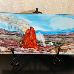 May include: A rectangular art piece depicting a landscape scene with a vibrant red rock formation and a white rock against a blue sky. The artwork is displayed on a black metal stand, showcasing the colorful abstract painting.