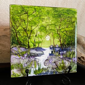 May include: A square tile featuring an alcohol ink painting of a lush green forest with a central white stream flowing through purple wildflowers and rocks. The artwork displays vibrant shades of lime and emerald green with a small white moon.