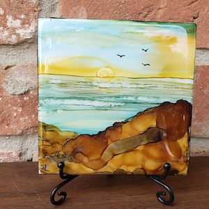 May include: A square glass tile with a sunset over the ocean scene. The tile is painted with shades of blue, green, yellow, and brown. There are three small black birds flying in the sky. The tile is on a black metal stand.