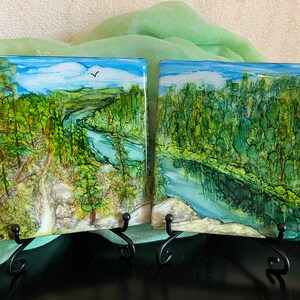 May include: Two square paintings on easels, each depicting a green forest scene with a river winding through it. The painting on the left has a bird flying in the sky. Both paintings have a blue sky with white clouds.
