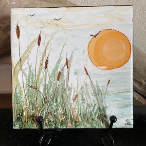 May include: Square artwork depicting a serene wetland scene. The painting features tall green reeds, brown cattails, and a bright orange sun. Two birds fly in the sky above the water. The artwork is displayed on a black metal stand.