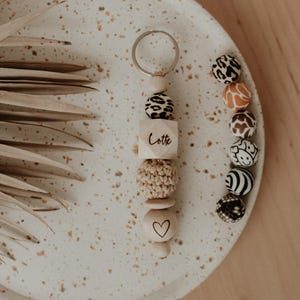 Può includere: Un portachiavi in legno con un ciondolo a forma di cuore e una perla di legno con la parola "Lotte" incisa. Il portachiavi è decorato con perline colorate con motivi di stampa animalier.