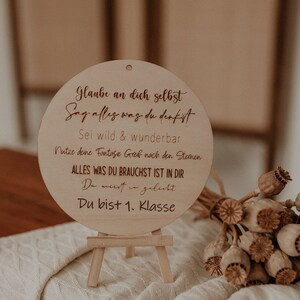 Puede incluir: Un letrero redondo de madera con un caballete. El letrero tiene un mensaje motivacional en alemán: "Glaube an dich selbst, Sag alles was du denkst, Sei wild & wunderbar, Nutze deine Fantasie greif nach den Sternen, Alles was du brauchst ist in dir, Du musst in geliebt, Du bist 1. Klasse".