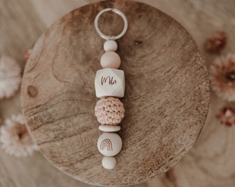 May include: A wooden bead keychain with a pink and white color scheme. The keychain features a wooden bead with the name "Mia" engraved on it, a crocheted bead, and a wooden bead with a rainbow design.