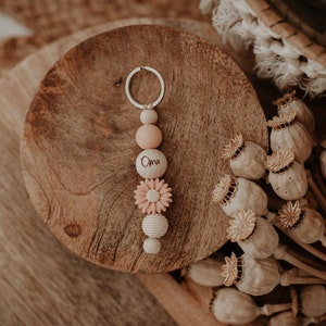 Può includere: Un portachiavi di perline di legno con un ciondolo a forma di fiore rosa e bianco e una perlina personalizzata che dice "Oma". Il portachiavi è su una superficie di legno con baccelli di semi di papavero secchi.