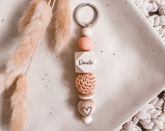 May include: A wooden keychain with a pink and beige color scheme. The keychain features a wooden bead with the name "Daniela" engraved on it, a crocheted bead, a wooden bead with a heart carved into it, and a wooden bead with a small ring attached. The keychain is on a white plate with a beige background.