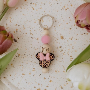 Könnte beinhalten: Ein Minnie-Mouse-Schlüsselanhänger mit Leopardenmuster und einer rosa Schleife. Der Schlüsselanhänger hat Holzperlen und einen silbernen Schlüsselring. Das Wort "Miri" ist auf dem Schlüsselanhänger geschrieben. Rosa und weiße Tulpen umgeben den Schlüsselanhänger.