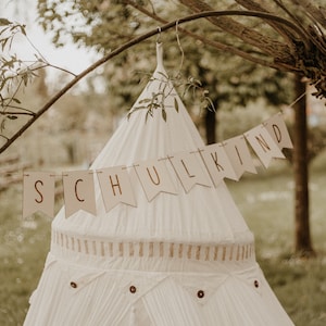 May include: A white teepee tent with a brown banner that says "SCHULKIND" hanging from a tree branch.