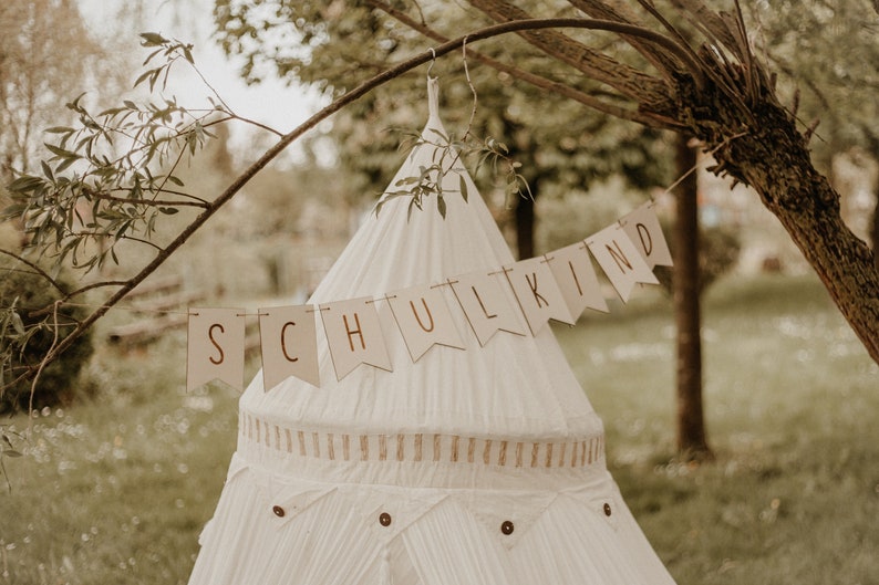May include: A white teepee tent with a brown banner that says "SCHULKIND" hanging from a tree branch.
