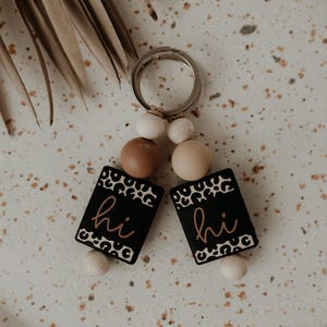 Puede incluir: Un llavero con dos colgantes rectangulares negros con borde de estampado de leopardo y la palabra "hi" en color cobre. El llavero tiene cuentas de madera y un anillo plateado.