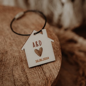 Può includere: Un portachiavi in legno bianco a forma di casa con un cinturino in pelle nera. Il portachiavi è inciso con "A&D" e un cuore, e la data "16.06.2014".