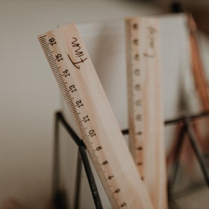 Puede incluir: Dos reglas de madera con los nombres "Finn" y "Henry" grabados en ellas. Las reglas est&aacute;n en un soporte de metal negro.