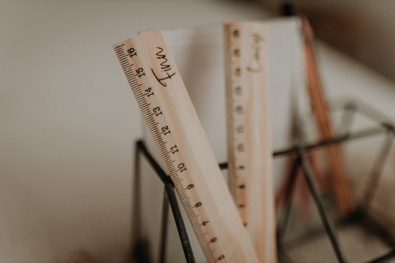 Puede incluir: Dos reglas de madera con los nombres "Finn" y "Henry" grabados en ellas. Las reglas est&aacute;n en un soporte de metal negro.