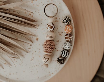 May include: A wooden bead keychain with a heart charm and the name "Milla" engraved on a bead. The keychain is decorated with animal print beads in brown, black, and white.
