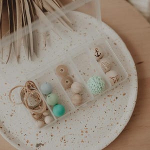 Könnte beinhalten: Eine durchsichtige Kunststoff-Sortierbox mit verschiedenen Bastelperlen. Die Fächer enthalten hellblaue, mintgrüne und türkisfarbene Silikonperlen, naturbelassene Holzperlen, eine gehäkelte mintfarbene Perle sowie Holzperlen mit Anker- und Regenbogenmotiven.