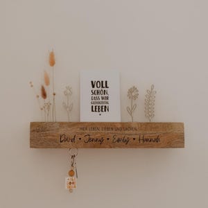 Könnte beinhalten: Ein Holzregal mit den eingravierten Namen "David, Jenny, Emily, Hannah". Das Regal trägt auch die Worte "Hier Leben, Lieben und Lachen" und eine Karte mit dem Text "VOLL SCHON, DASS WIR GLEICHZEITIG LEBEN". Ein Schlüsselanhänger hängt darunter.
