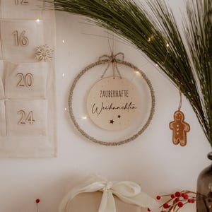 Könnte beinhalten: Ein kreisförmiger, mit silbernem Lametta umwickelter Dekoreifen, der in der Mitte eine Holzscheibe mit dem Text Zauberhafte Weihnachten und kleinen Sternsymbolen trägt. Der Reifen ist an einer silbernen Schnur befestigt und hat eine kleine Schleife.