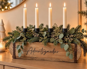 May include: A rectangular wooden box with metal accents featuring the text Adventszauber in black script. The box holds four black candle holders with lit off-white taper candles, surrounded by green pine branches and eucalyptus leaves.