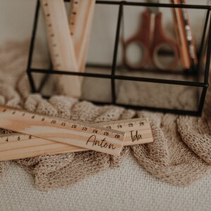 Puede incluir: Dos reglas de madera con los nombres "Anton" e "Ida" grabados en ellas. Las reglas est&aacute;n sobre una tela de punto beige.