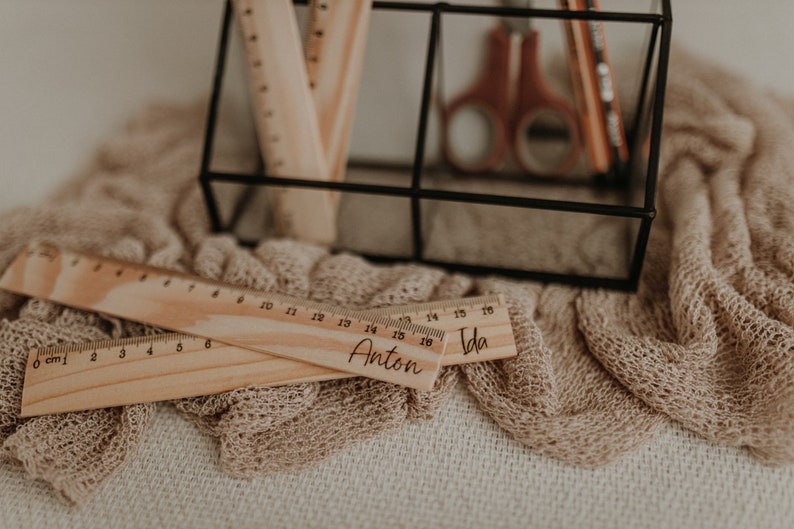 Puede incluir: Dos reglas de madera con los nombres "Anton" e "Ida" grabados en ellas. Las reglas est&aacute;n sobre una tela de punto beige.
