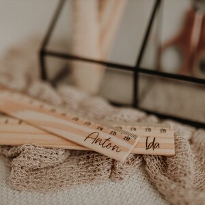 Puede incluir: Dos reglas de madera con los nombres "Anton" e "Ida" grabados en ellas. Las reglas est&aacute;n sobre una superficie de tela beige.