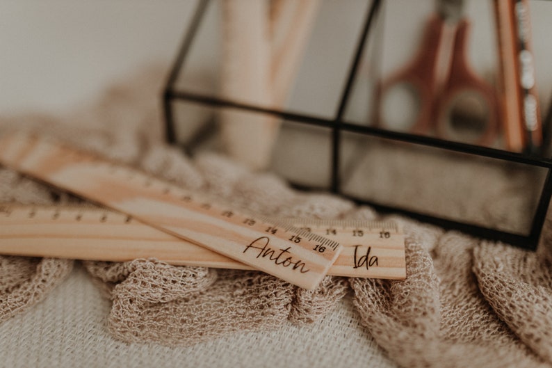Puede incluir: Dos reglas de madera con los nombres "Anton" e "Ida" grabados en ellas. Las reglas est&aacute;n sobre una superficie de tela beige.