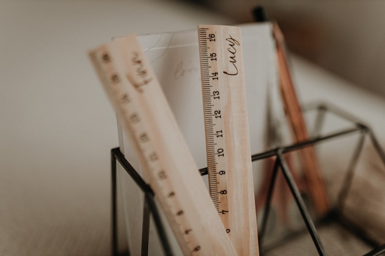 Puede incluir: Dos reglas de madera con marcas negras, una con el nombre "Lucy" grabado, est&aacute;n de pie en un soporte geom&eacute;trico de metal negro.
