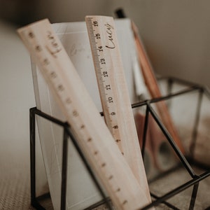 Puede incluir: Dos reglas de madera con marcas en pulgadas, una con el nombre "Lucy" inscrito en ella, se muestran en un soporte geom&eacute;trico de metal negro. El soporte est&aacute; sobre una superficie de tela beige.