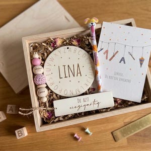 Op de afbeelding: Een houten geschenkdoos met een gepersonaliseerde sleutelhanger, een rond houten bord met de naam "LINA", een pen met een hondenopzetstuk en een wenskaart. De kaart leest "Juhuuu. Du bist ein Schulkind".