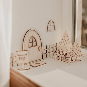 Peut inclure: Maison miniature en bois avec une porte, une fenêtre, une clôture, une boîte aux lettres et deux sapins de Noël. La maison est en bois clair et a un toit brun. La boîte aux lettres indique "Post".