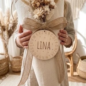 Puede incluir: Un ramo de flores y follaje secos atado con una cinta de arpillera. Un disco de madera con el nombre "LINA" está sujeto a la cinta. El ramo está envuelto en una tela color crema.