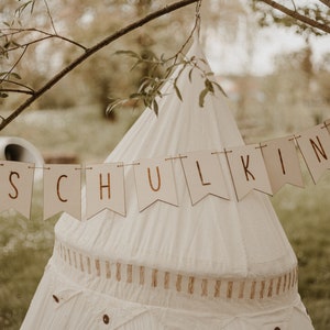 May include: A white teepee tent with a banner that reads "SCHULKIND" hanging above it. The banner is made of brown paper with gold lettering. The teepee is set up outdoors in a grassy area.