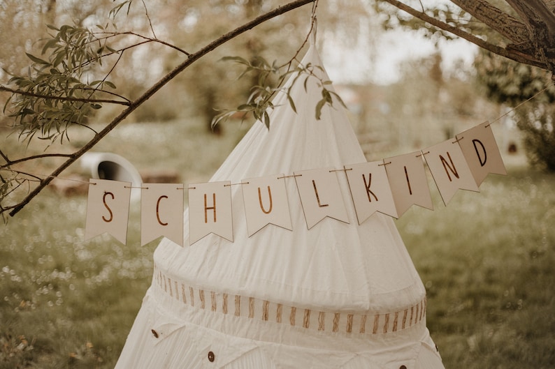 May include: A white teepee tent with a banner that reads "SCHULKIND" hanging above it. The banner is made of brown paper with gold lettering. The teepee is set up outdoors in a grassy area.