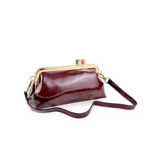 May include: A deep burgundy clutch bag with a glossy finish, featuring a gold-toned metal frame closure with a square and spherical clasp. The bag includes a matching detachable shoulder strap attached to gold-toned hardware on both sides.