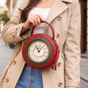 May include: A red circular handbag featuring a clock face design. The bag has a gold-coloured frame and a handle. The background shows a cobbled street and buildings.
