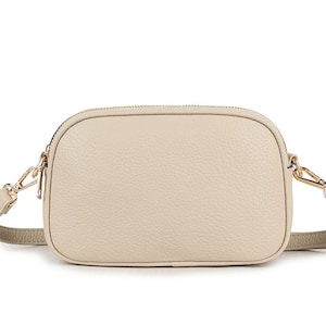 Puede incluir: Un bolso bandolera beige con forma rectangular y cierre de cremallera. El bolso tiene una superficie texturizada y herrajes dorados, incluyendo los enganches de la correa ajustable. La correa también es beige.