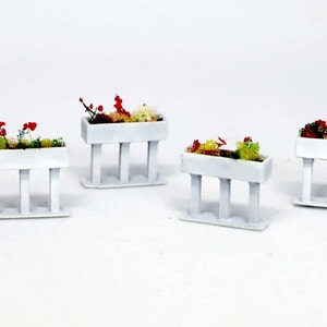 May include: Four white planter boxes with red and green flowers on white stands. The planter boxes are rectangular and have a simple design.