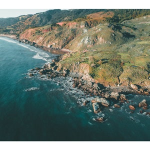 May include: Aerial view of a rocky coastline with turquoise water crashing against the shore. Lush green hills and cliffs rise up in the background.