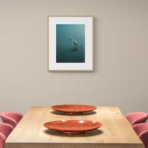Op de afbeelding: Een houten eettafel met twee rode stoelen en twee rode borden. Een ingelijst foto van een witte vogel in het water hangt aan de muur achter de tafel.