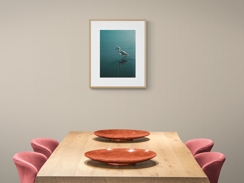 Op de afbeelding: Een houten eettafel met twee rode stoelen en twee rode borden. Een ingelijst foto van een witte vogel in het water hangt aan de muur achter de tafel.