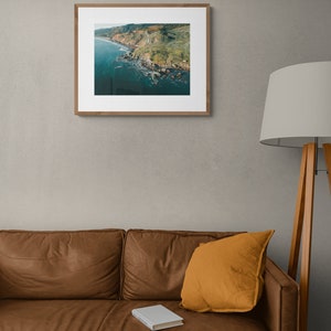 May include: An aerial photograph of a rocky coastline with green hills and blue water. The photo is framed in a light wood frame and hanging on a gray wall.