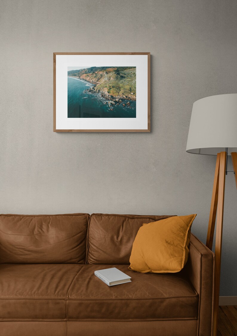 May include: An aerial photograph of a rocky coastline with green hills and blue water. The photo is framed in a light wood frame and hanging on a gray wall.